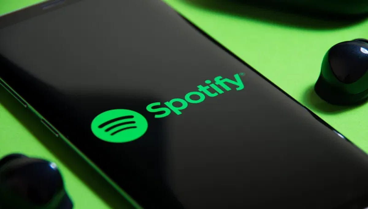 نمای نزدیک از گوشی هوشمند با لوگوی Spotify روی صفحه، در کنار هدفون‌های بی‌سیم، که به قابلیت‌های تعاملی و بازی‌محور اسپاتیفای مانند Heardle و تجربه شنیداری دیجیتال اشاره دارد.
