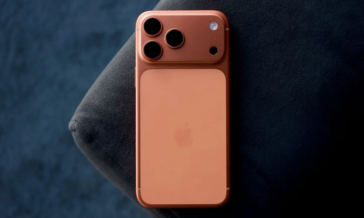 نمای پشت یک آیفون با بدنه فلزی به رنگ نارنجی–مسی و ماژول دوربین سه‌گانه، قرارگرفته روی سطح پارچه‌ای تیره؛ طراحی مینیمال اپل با تمرکز بر متریال و چیدمان دوربین‌ها