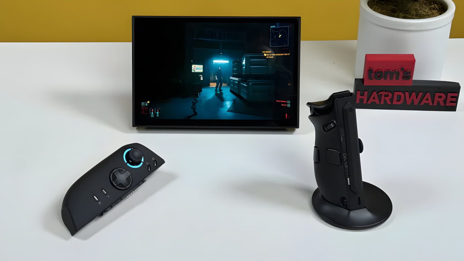ستاپ گیمینگ شامل یک نمایشگر کوچک با اجرای بازی Cyberpunk 2077، کنترلر دستی ماژولار در دو حالت افقی و عمودی، قرارگرفته روی میز سفید در کنار پایه نمایش با برند Tom’s Hardware.