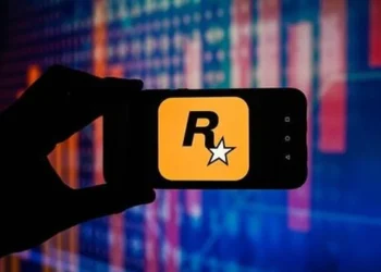 نمای نزدیک از گوشی هوشمند که در دست یک فرد نگه داشته شده و لوگوی زرد رنگ Rockstar Games با حرف R و ستاره سفید روی صفحه نمایش داده می‌شود؛ پس‌زمینه شامل افکت‌های نورانی و خطوط گرافیکی شبیه نمودارهای دیجیتال