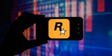 نمای نزدیک از گوشی هوشمند که در دست یک فرد نگه داشته شده و لوگوی زرد رنگ Rockstar Games با حرف R و ستاره سفید روی صفحه نمایش داده میشود؛ پسزمینه شامل افکتهای نورانی و خطوط گرافیکی شبیه نمودارهای دیجیتال