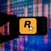 نمای نزدیک از گوشی هوشمند که در دست یک فرد نگه داشته شده و لوگوی زرد رنگ Rockstar Games با حرف R و ستاره سفید روی صفحه نمایش داده می‌شود؛ پس‌زمینه شامل افکت‌های نورانی و خطوط گرافیکی شبیه نمودارهای دیجیتال