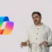 تصویر ارائه‌ای با پس‌زمینه سفید که در آن لوگوی رنگین‌کمانی Microsoft Copilot در سمت چپ دیده می‌شود و فردی در سمت راست در حال توضیح دادن با حرکات دست است؛ نمایی مفهومی از معرفی قابلیت‌های هوش مصنوعی کوپایلت مایکروسافت