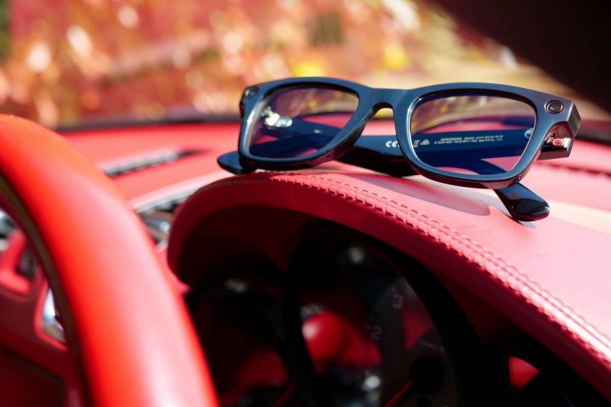 تصویر عینک هوشمند Ray‑Ban Stories با فریم مشکی و دوربین تعبیهشده در گوشه فریم که روی داشبورد چرمی قرمزرنگ یک خودرو قرار گرفته است