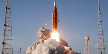 لحظه پرتاب یک موشک فضایی (آرتمیس ۲) از سکوی پرتاب؛ شعله‌های موتور و حجم بزرگی از دود سفید در اطراف پایگاه دیده می‌شود و موشک نارنجی‑سفید در حال اوج گرفتن به سمت آسمان آبی است