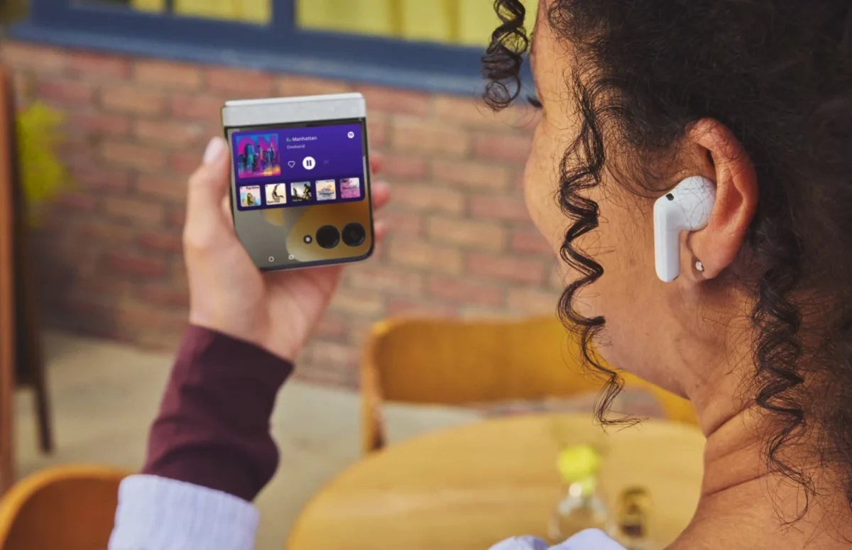 فردی در حال استفاده از گوشی تاشوی سامسونگ Galaxy Z Flip با نمایشگر بیرونی فعال و پخش موسیقی در محیط کافه؛ تمرکز بر تجربه کاربری و طراحی قابلتاشدن