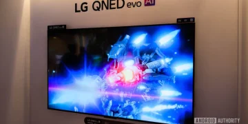 تلویزیون بزرگ LG QNED evo AI روی دیوار نمایشگاهی که صحنهای علمیتخیلی با نورهای آبی و قرمز را نمایش میدهد؛ واترمارک Android Authority در گوشه تصویر دیده میشود