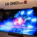 تلویزیون بزرگ LG QNED evo AI روی دیوار نمایشگاهی که صحنهای علمیتخیلی با نورهای آبی و قرمز را نمایش میدهد؛ واترمارک Android Authority در گوشه تصویر دیده میشود