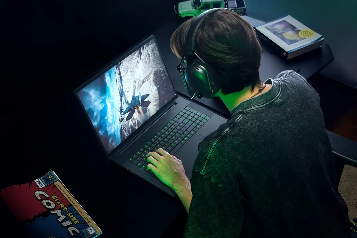 شخصی با هدست گیمینگ پشت لپ‌تاپ Razer Blade نشسته است. صفحه‌نمایش لپ‌تاپ تصویری از یک جت جنگنده در حال پرواز روی زمین برفی را نشان می‌دهد. نور سبزرنگ محیط و کیبورد RGB فضای گیمینگ را برجسته می‌کند. یک کتاب کمیک در سمت چپ میز دیده می‌شود.