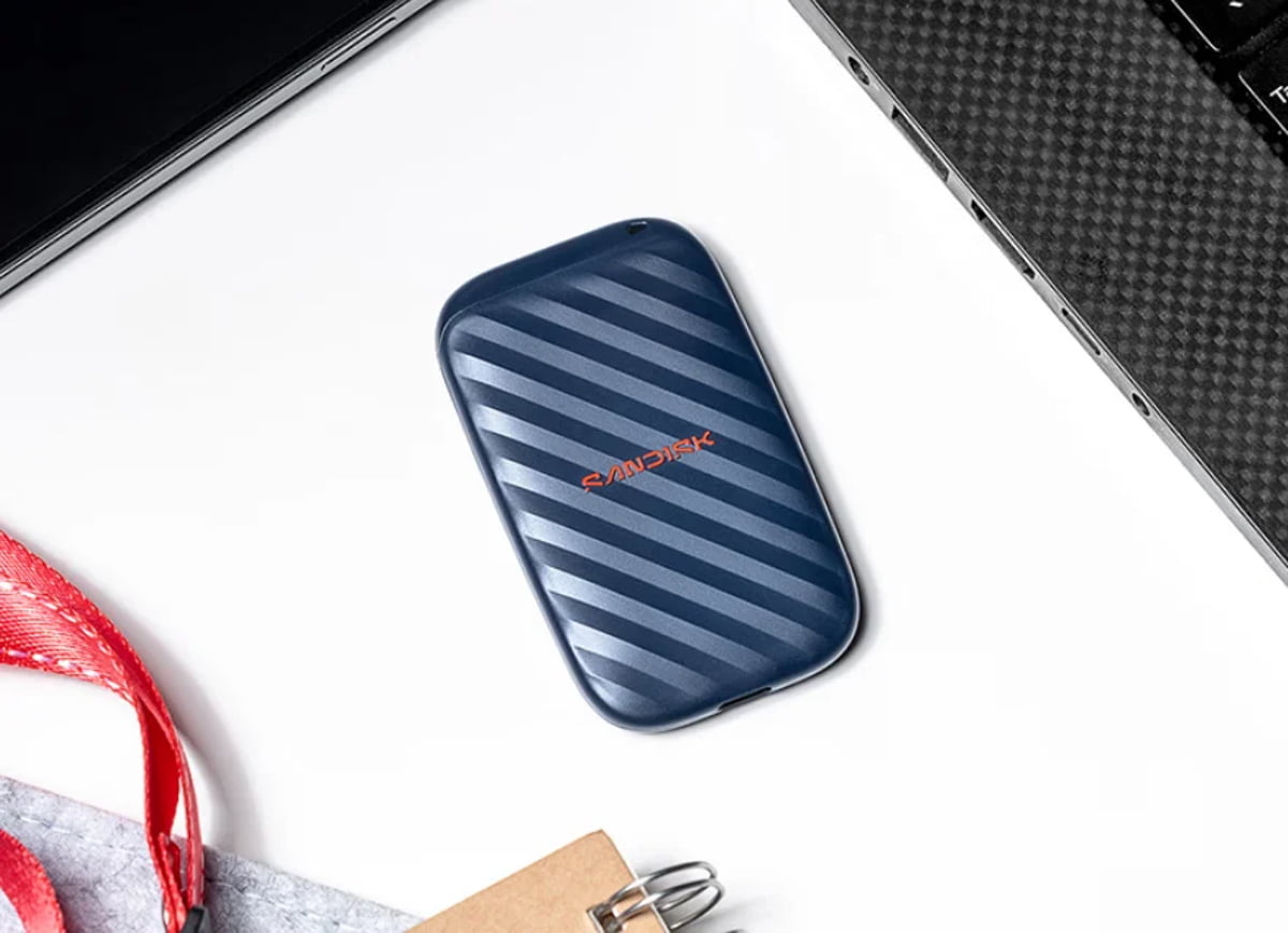 هارد اکسترنال قابل‌حمل SanDisk آبی‌رنگ با خطوط مورب که روی میز سفید کنار لپ‌تاپ و لوازم جانبی قرار گرفته است