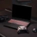 ستاپ گیمینگ شامل لپتاپ گیمینگ با کیبورد RGB قرمز، کنترلر Xbox، ماوس گیمینگ و هدست روی میز در محیط نورپردازی تیره.