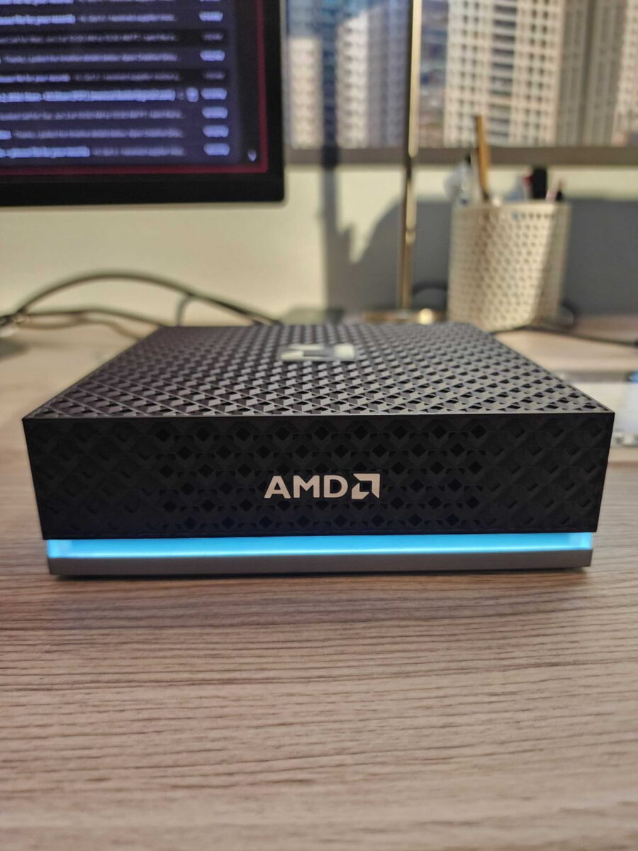 نمای نزدیک از یک مینی پیسی مشکی برند AMD با بدنه مشبک هندسی و نوار نورانی LED آبی در لبه پایینی. دستگاه روی یک میز چوبی قرار دارد و لوگوی نقرهای AMD در جلو دیده میشود