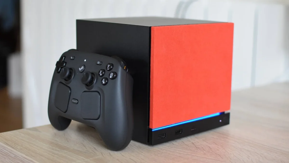 نمای نزدیک از کنسول Steam Machine با پنل جلویی نارنجی و نورپردازی آبی در کنار Steam Controller مشکی روی میز چوبی