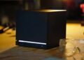 کنسول مشکیرنگ و مکعبیشکل استیم ماشین (Steam Machine) با نورپردازی LED خطی سفید در پایین، قرار گرفته روی میز کار در کنار عینک ایمنی و ابزارآلات