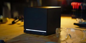 کنسول مشکی‌رنگ و مکعبی‌شکل استیم ماشین (Steam Machine) با نورپردازی LED خطی سفید در پایین، قرار گرفته روی میز کار در کنار عینک ایمنی و ابزارآلات