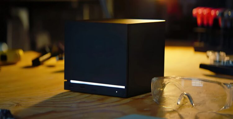کنسول مشکیرنگ و مکعبیشکل استیم ماشین (Steam Machine) با نورپردازی LED خطی سفید در پایین، قرار گرفته روی میز کار در کنار عینک ایمنی و ابزارآلات