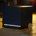 کنسول مشکی‌رنگ و مکعبی‌شکل استیم ماشین (Steam Machine) با نورپردازی LED خطی سفید در پایین، قرار گرفته روی میز کار در کنار عینک ایمنی و ابزارآلات