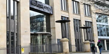 نمای بیرونی ساختمان مدرن و سنگی استودیو Rockstar North در ادینبرگ اسکاتلند با لوگوی بزرگ راکاستار نورث بالای ورودی چرخشی. عابران در حال عبور از مقابل نردههای حفاظتی ساختمان هستند
