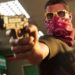 نمای نزدیک از شخصیت جیسون در GTA VI که یک اسلحه کمری به سمت دوربین گرفته و صورت خود را با یک دستمال گردن (باندانا) قرمز و عینک آفتابی زرد پوشانده است