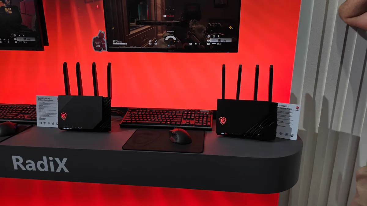 غرفه MSI در نمایشگاه با پس‌زمینه قرمز و میز RadiX؛ دو روتر گیمینگ Wi‑Fi با چند آنتن کنار کیبورد و ماوس و نمایش گیم روی مانیتور