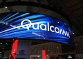 تابلوی بزرگ Qualcomm روی نمایشگر LED منحنی در یک سالن نمایشگاهی با نورپردازی آبی؛ نمای غرفه و سقف سالن در پسزمینه