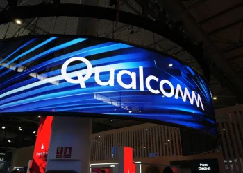 تابلوی بزرگ Qualcomm روی نمایشگر LED منحنی در یک سالن نمایشگاهی با نورپردازی آبی؛ نمای غرفه و سقف سالن در پس‌زمینه