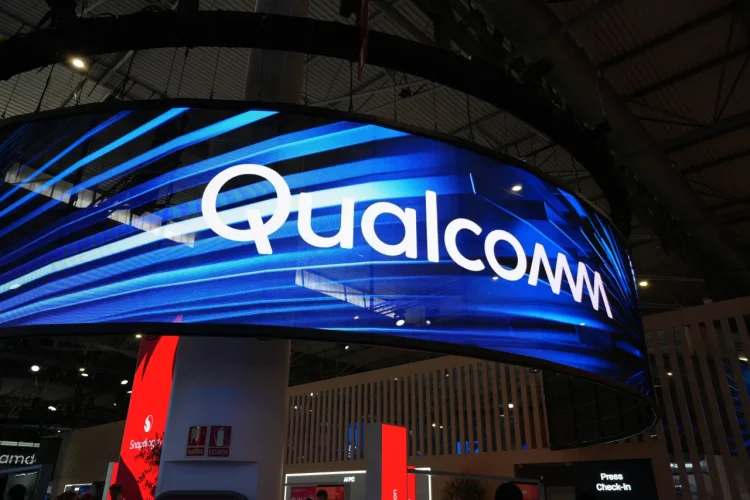 تابلوی بزرگ Qualcomm روی نمایشگر LED منحنی در یک سالن نمایشگاهی با نورپردازی آبی؛ نمای غرفه و سقف سالن در پس‌زمینه