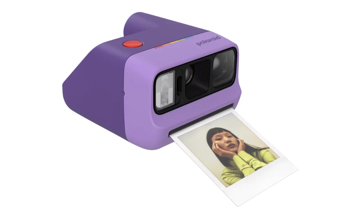 دوربین فوری Polaroid Go Generation 3 به رنگ بنفش در حال چاپ یک عکس فوری؛ نمای نزدیک از طراحی جمع‌وجور، لنز و فلش دستگاه.