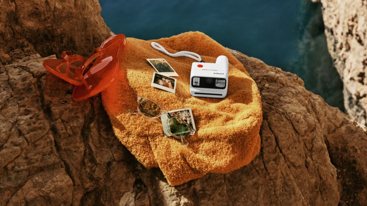 دوربین Polaroid Go Generation 3 در کنار عکس‌های چاپ‌شده، عینک آفتابی و لوازم سفر روی صخره‌ای در فضای باز؛ مناسب برای ثبت لحظات سفر و ماجراجویی