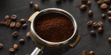 پودر قهوه آسیابشده داخل پرتافیلتر فلزی در کنار دانههای قهوه روی سطح تیره برای نمایش آمادهسازی اسپرسو.