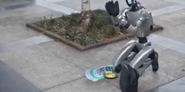 ربات انسان‌نمای Unitree G1 در خیابانی در شهر چنگدو چین روی زمین زانو زده و مقابل خود یک کد QR و ظرف دریافت کمک قرار داده است؛ یکی از رهگذران نیز در حال پرداخت پول و تعامل با این ربات است.