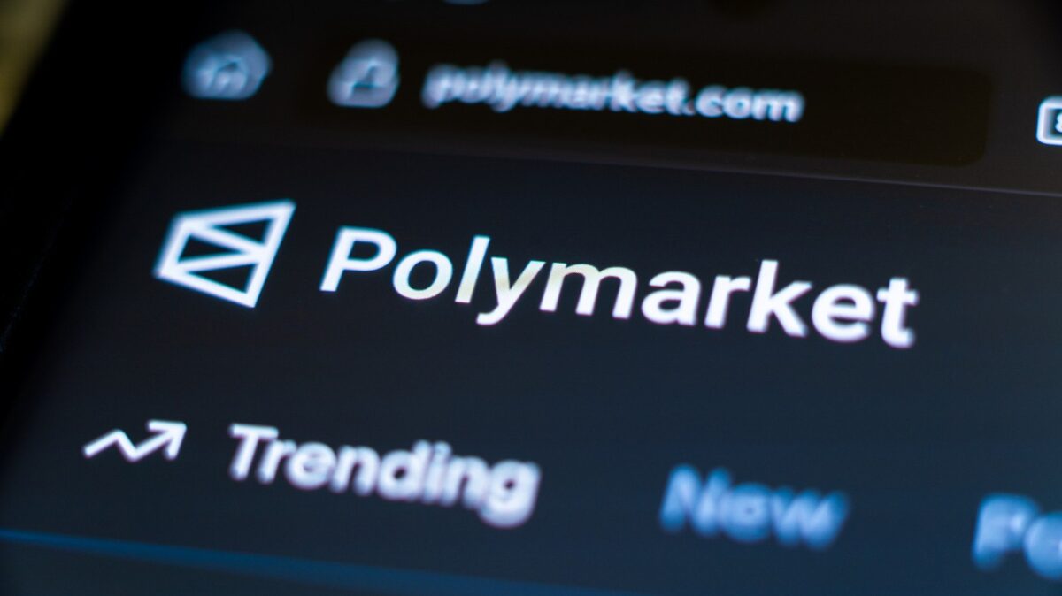 لوگوی Polymarket روی نمایشگر یک گوشی هوشمند در مقابل پسزمینه آبی با نشان این پلتفرم؛ تصویری نمادین از بازارهای پیشبینی و شرطبندی آنلاین.