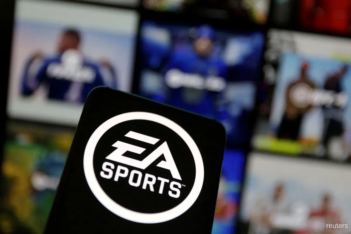 لوگوی سفید و مشکی EA Sports روی صفحه یک تلفن همراه، با تصاویر محو جلد بازیهای ورزشی در پسزمینه