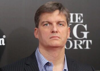 پرتره کلوزآپ از مایکل بری (Michael Burry)، سرمایه‌گذار مشهور و مدیر صندوق تامین سرمایه، با کت و شلوار راه راه و پیراهن آبی، در حال شرکت در یک رویداد تبلیغاتی فیلم The Big Short با پس‌زمینه برند Heineken
