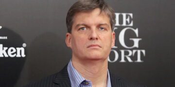 پرتره کلوزآپ از مایکل بری (Michael Burry)، سرمایه‌گذار مشهور و مدیر صندوق تامین سرمایه، با کت و شلوار راه راه و پیراهن آبی، در حال شرکت در یک رویداد تبلیغاتی فیلم The Big Short با پس‌زمینه برند Heineken