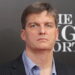 پرتره کلوزآپ از مایکل بری (Michael Burry)، سرمایهگذار مشهور و مدیر صندوق تامین سرمایه، با کت و شلوار راه راه و پیراهن آبی، در حال شرکت در یک رویداد تبلیغاتی فیلم The Big Short با پسزمینه برند Heineken