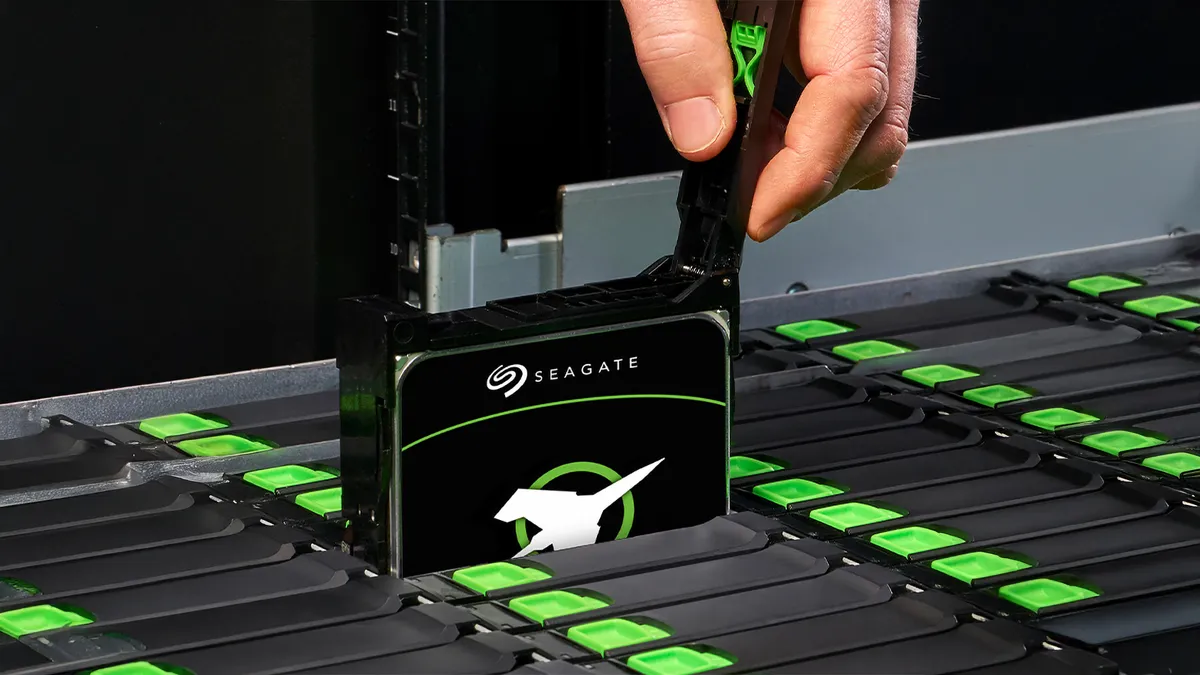 نمای نزدیک از دست یک تکنسین در حال جای‌گذاری یک هارد دیسک سازمانی Seagate Exos درون یک رک سرور پرظرفیت. طراحی ماژولار کیس با زبانه سبز رنگ و چیدمان منظم محفظه‌های هارد در فضای تاریک مرکز داده، نشان‌دهنده محیط‌های عملیاتی سرورهای صنعتی است.