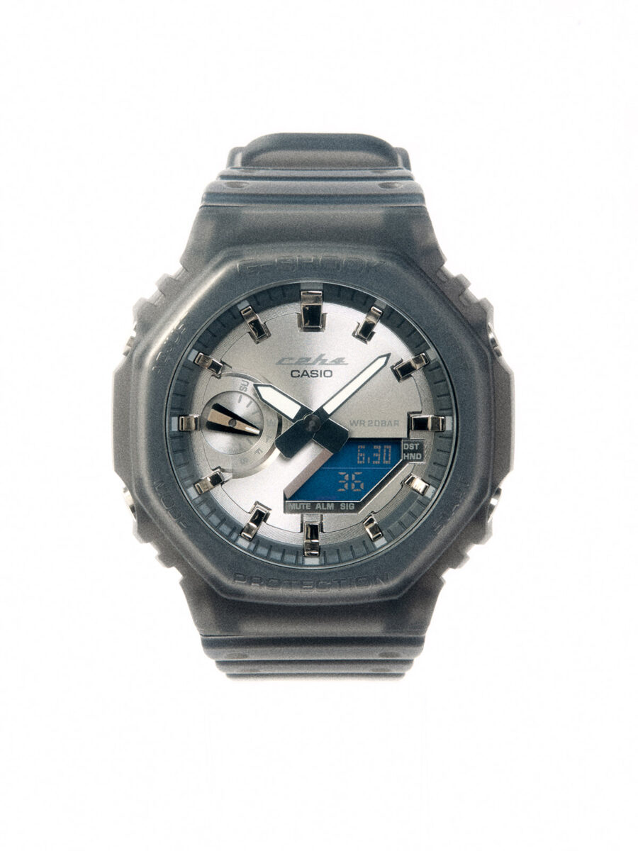 تصویر نمای نزدیک و باکیفیت از صفحه ساعت مچی Casio G-Shock مدل GA-2100 همکاری با C2H4؛ بدنه دودیرنگ و نیمهشفاف با عقربههای سفید و صفحه نمایش دیجیتال آبیرنگ در پسزمینه سفید.