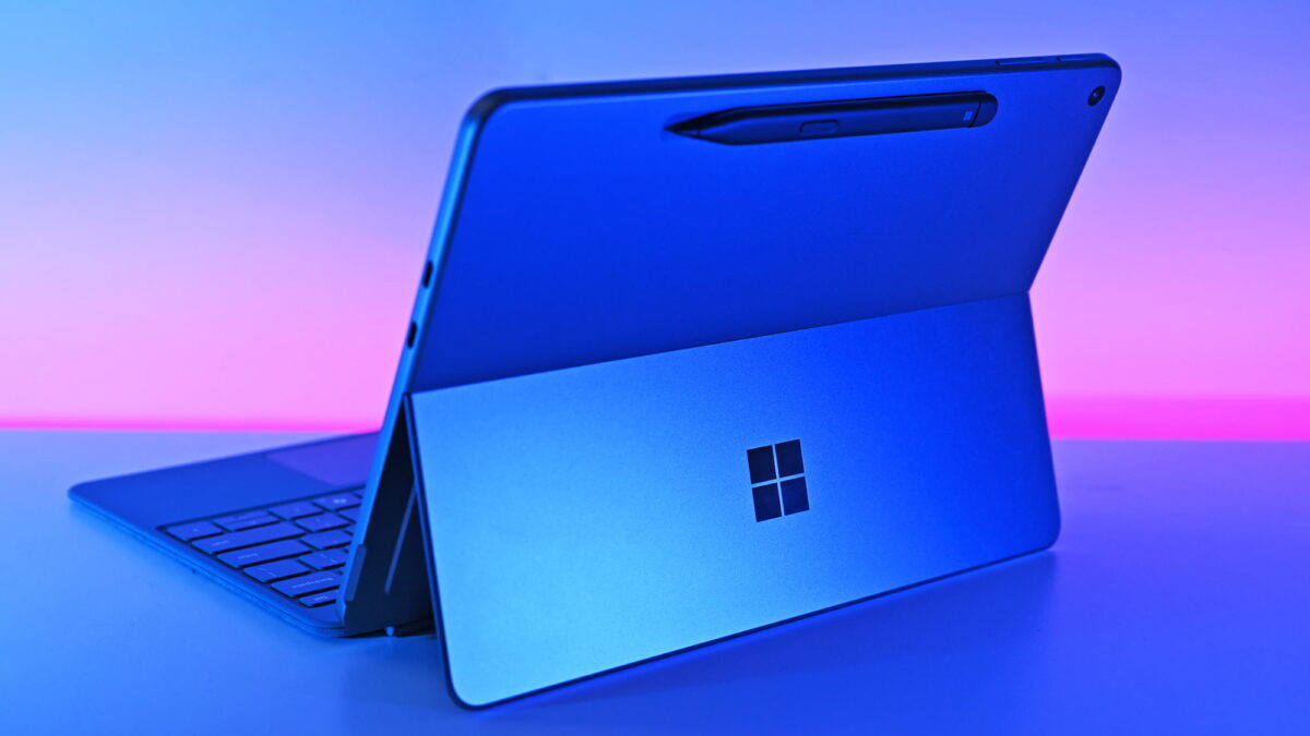 نمای سه‌چهارم از پشت یک تبلت آبی‌رنگ Microsoft Surface Pro با پایه باز، کیبورد متصل و قلم Surface؛ لوگوی مایکروسافت روی پایه دیده می‌شود و صحنه با نور نئونی آبی و صورتی روشن شده است.