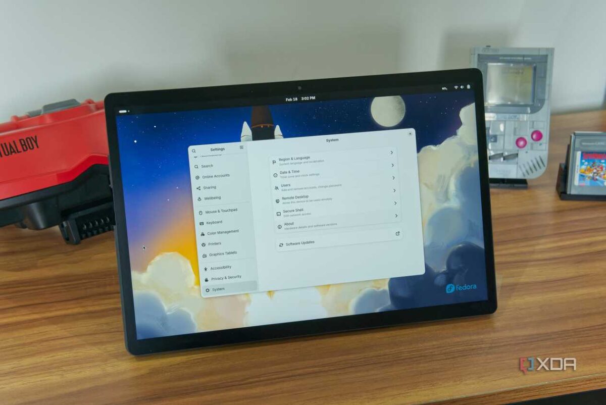 تبلتی روی میز چوبی که رابط دسکتاپ Fedora Linux و تنظیمات GNOME را نمایش میدهد، با کنسولهای بازی قدیمی مانند Virtual Boy و Game Boy در پسزمینه.
