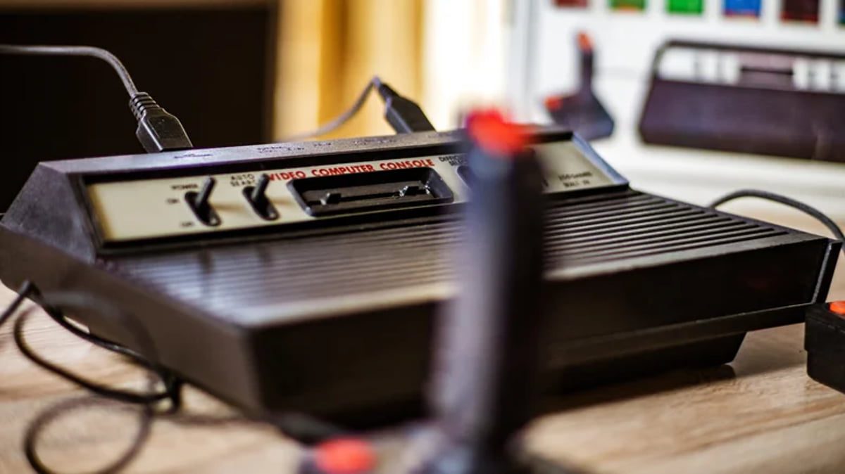 نمای زاویه‌دار از کنسول بازی کلاسیک آتاری (Atari Video Computer Console) به همراه جوی‌استیک؛ کلیدهای پاور و تنظیمات کنسول در پنل جلویی به وضوح دیده می‌شوند