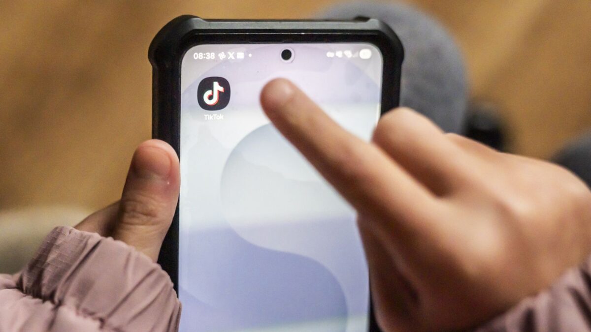 نمای نزدیک از دست یک کودک یا نوجوان در حال لمس آیکون اپلیکیشن تیک‌تاک (TikTok) روی صفحه نمایش یک گوشی اندرویدی؛ گوشی دارای یک قاب محافظ مشکی ضخیم است و محیط با نوری گرم و خانگی دیده می‌شود