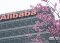 نمای زاویهدار از بالای ساختمان شرکت علیبابا با تابلوی بزرگ و نارنجیرنگ Alibaba در بالای نما، در حالی که شاخههای پر از شکوفههای صورتی درختان در پیشزمینه تصویر قرار دارند.