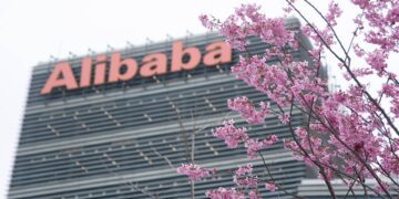 نمای زاویهدار از بالای ساختمان شرکت علیبابا با تابلوی بزرگ و نارنجیرنگ Alibaba در بالای نما، در حالی که شاخههای پر از شکوفههای صورتی درختان در پیشزمینه تصویر قرار دارند.