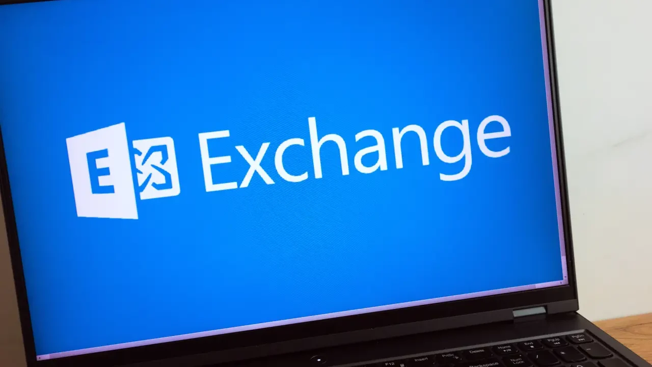 نمای نزدیک لپتاپ روی میز که لوگوی Microsoft Exchange را روی پسزمینه آبی نمایش میدهد.
