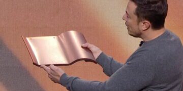 ایلان ماسک در یک برنامه یا مراسم نمایشی، بخشی از یک پنل خورشیدی فلزی و خمیده را با هر دو دست باز کرده و بررسی میکند.