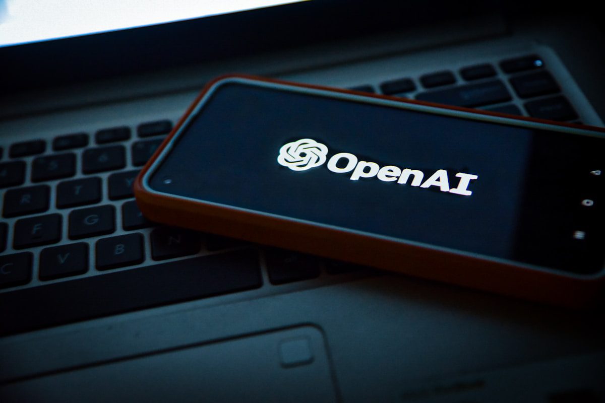 گوشی هوشمندی با لوگوی درخشان OpenAI که روی کیبورد یک لپتاپ در محیطی با نور کم قرار گرفته است.