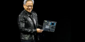 جنسن هوانگ (Jensen Huang)، مدیرعامل انویدیا، با کاپشن چرم مشکی روی صحنه در حال لبخند زدن و در دست داشتن یک برد مدار الکترونیکی پیشرفته و مادربورد سرور هوش مصنوعی.