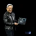 جنسن هوانگ (Jensen Huang)، مدیرعامل انویدیا، با کاپشن چرم مشکی روی صحنه در حال لبخند زدن و در دست داشتن یک برد مدار الکترونیکی پیشرفته و مادربورد سرور هوش مصنوعی.