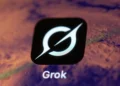 نمای نزدیک از لوگوی سفید Grok روی یک مربع مشکی، در برابر پس‌زمینه‌ای تار و بنفش‌رنگ که بخشی از رابط کاربری یا نمایشگر را نشان می‌دهد.